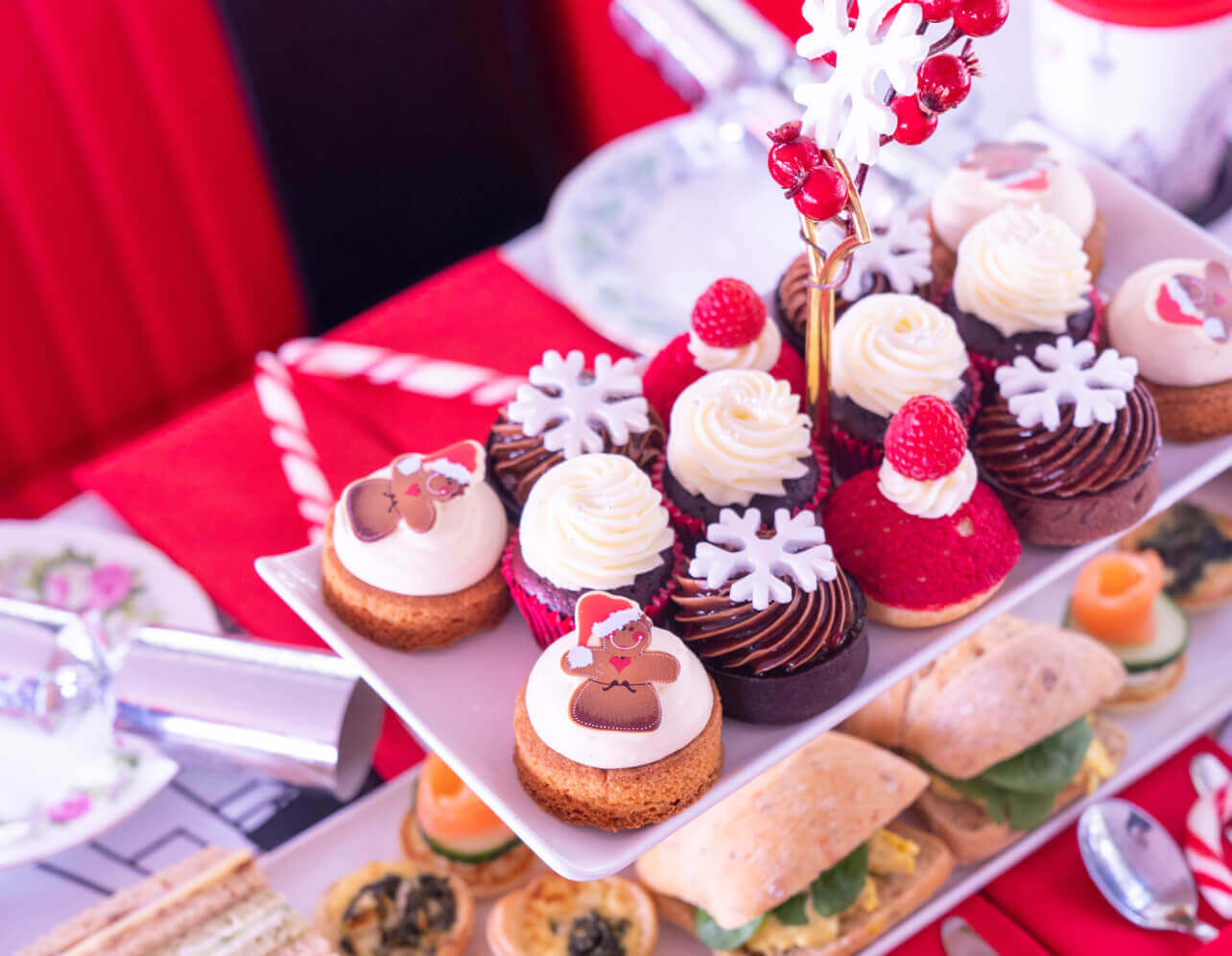 Stand with festive themed cakes and pastries and bottom stand with savouries, atop a table with Christmas crackers  and other Christmas decorations